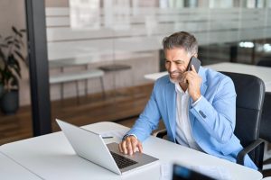 Homme d’affaires souriant en conversation téléphonique tout en travaillant sur un ordinateur portable dans un bureau moderne.
